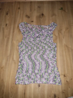 Lila och grön virkad ärmlös topp - Handvirkad topp i lila, grönt och vitt med vågigt mönster. Modellen är ärmlös och har en rak passform. Perfekt för dig som gillar unika plagg med retrovibbar. Materialet är stickat garn som ger en mysig känsla. Virkad av mamma, därför lite dyrare🥰