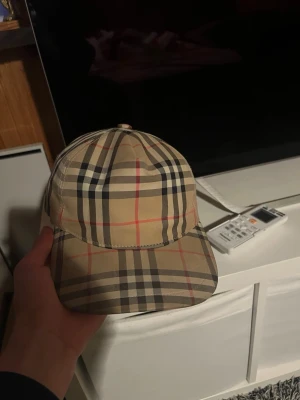 Burberry beige rutmönstrad keps - Säljer en klassisk beige keps från Burberry med det ikoniska rutmönstret i svart, vitt och rött. Kepsen har justerbar snapback och en svart patch med texten 'Burberry London England' bak. Perfekt för dig som gillar stilrena accessoarer.