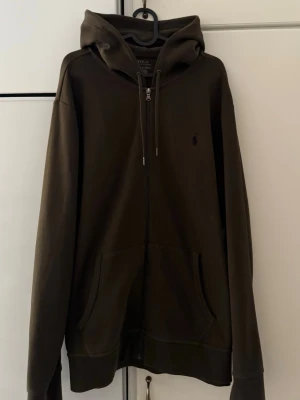 Mörkgrön hoodie från Polo Ralph Lauren - Snygg mörkgrön hoodie från Polo Ralph Lauren med dragkedja framtill, två stora fickor och klassisk huva med dragsko. Liten broderad logga på bröstet. Tillverkad i mjuk bomullsblandning som känns skön mot huden. Perfekt för en avslappnad och stilren look.