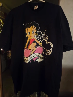 Svart Betty Boop t-shirt - Cool svart t-shirt med stor Betty Boop-tryck på framsidan. Motivet visar Betty Boop sittandes på ett rosa hjärta med texten 'LOVE' och 'BETTY BOOP' i banderoller. Klassisk passform och rund hals, perfekt för dig som gillar retro och popkultur.