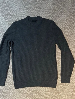 Mörkgrå stickad tröja J.Lindeberg - Snygg mörkgrå stickad tröja från J.Lindeberg med rund halsringning och ribbade muddar. Tröjan har ett diskret ribbstickat mönster och är perfekt för lager på lager. Klassisk och stilren design som passar till det mesta.