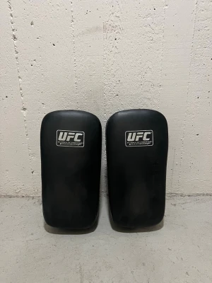 UFC Mittsar för Thaiboxning/Kampsport - Ett par svarta thaiboxnings sparkmittsar från UFC. Tillverkade i slitstarkt konstläder med rektangulär form, rejäla handtag och justerbara remmar på baksidan för stabilt grepp. Tydlig UFC-logga på fram- och baksida. Använda bara 1 gång, legat i garaget sedan dess. Perfekta för kampsportsträning! 