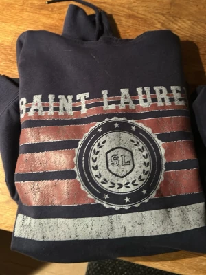 Mörkblå Saint Laurent hoodie - Mörkblå hoodie med tryck i rött, vitt och grått framtill. Stor logga med texten 'SAINT LAURENT' och emblem med 'SL' på bröstet. Klassisk huva med dragsko och magficka. Perfekt för en chill och avslappnad stil.
