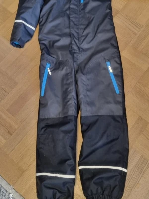 Mörkgrå vinteroverall från Reima  - Mörkgrå vinteroverall med turkos dragkedjor och reflexdetaljer vid bensluten. Tillverkade i slitstark syntet, perfekta för kalla vinterdagar. Två fickor framtill med blå detaljer och elastiska muddar vid benslut. Storlek 140. Ingen sköljmedel använt på den. Namn skrivit på etiketten i nacken.