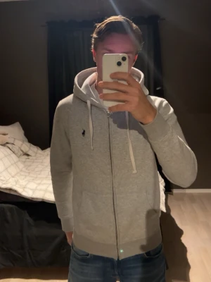 Polo Ralph Lauren zip hoodie - Grå hoodie från Polo Ralph Lauren med dragkedja och vit snörning i huvan. Klassisk logga broderad på bröstet. Perfekt för en avslappnad och stilren look. Sparsamt använd (som ny). Hör av dig om du undrar något eller vill ha fler bilder så svarar jag så snabbt som möjligt!          Bvsa!