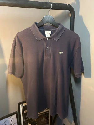 Marinblå piké från Lacoste - Klassisk marinblå pikétröja från Lacoste med korta ärmar och den ikoniska krokodilloggan på bröstet. Tröjan har krage, knappar framtill och är gjord i mjuk bomull som känns skön mot huden. Perfekt för en clean och stilren look.