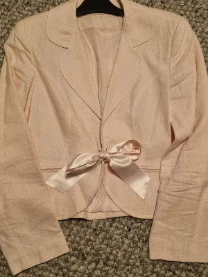 Ljusrosa linne  kavaj med sidenrosett - Snygg ljusrosa linne kavaj med klassisk krage och lång ärm. Kavajen har en elegant sidenrosett i midjan som ger en feminin touch. Perfekt för dig som vill ha en chic och stilren look. Köpt i Storbritannien. 