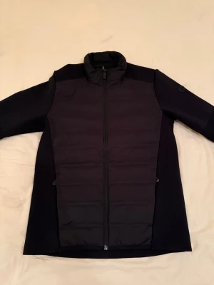 Sail Racing Cardigan - Stilren svart cardigan jacka med quiltad front och hel dragkedja. Jackan har långärmade stickade partier på ärmar och sidor, samt hög krage för extra värme. Perfekt för lager-på-lager och enkel att matcha med olika outfits. Köpt på Collection of Brands i Sollentuna Centrum. Nypris: 3000 kr