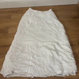 Vit långkjol från Stradivarius - Säljer en vit långkjol från Stradivarius med snyggt rynkad struktur. Kjolen har en luftig och flowy passform, perfekt för varma dagar. Tillverkad i ett lätt material som ger en avslappnad och trendig look.