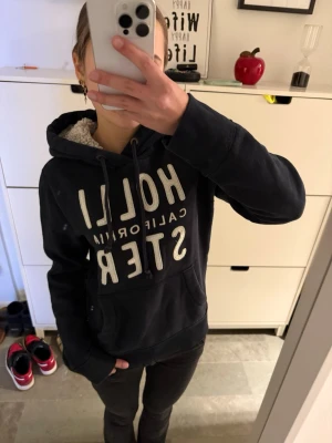 Fodrad mörkblå hoodie Hollister - Mysig svart hoodie från Hollister med stor vit logga på bröstet'. Hoodien har dragsko i huvan och mjukt, fluffigt foder på insidan av huvan. Perfekt för dig som gillar en avslappnad och trendig stil.