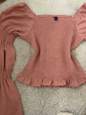 Rosa smockad blus med volangärm - Supersöt rosa blus med smockad kropp och puffiga långärmar. Toppen har volangkant nedtill och vid ärmslut, samt en offshoulder-design som ger en trendig look. Perfekt för dig som gillar romantiska detaljer och en feminin stil.