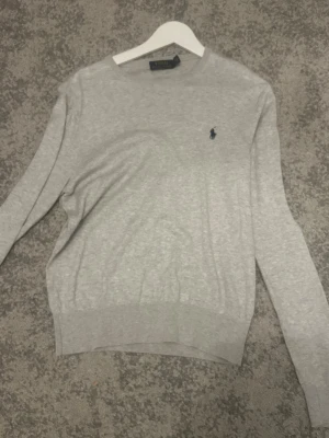 Grå kashmir och bomullströja från Polo Ralph Lauren - Snygg ljusgrå tröja från Polo Ralph Lauren med rund halsringning och diskret broderad logga på bröstet. Tröjan har långa ärmar och ribbade muddar vid ärmslut och nederkant. Perfekt för en stilren och klassisk look. Kashmir och bomull