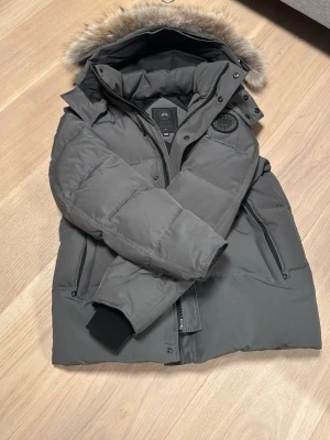 Grå Canada Goose pufferjacka med päls - Säljer en grå pufferjacka från Canada Goose i storlek M. Jackan har en fluffig huva med päls, svarta muddar vid ärmslut och dragkedja framtill. Märket syns tydligt på armen och jackan har en clean, modern look som är perfekt för kalla dagar. Jackan är nästan i nyskick. Allting original medföljer.