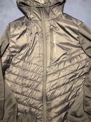 Grå vindjacka Sail Racing med huva - Snygg grå vindjacka från Sail Racing med huva och hel dragkedja framtill. Jackan har quiltade partier på fram- och baksidan samt släta ärmar. Perfekt för blåsiga dagar och har en sportig look med diskret logga på bröstet.