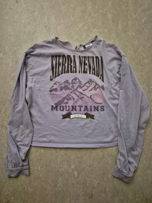 Tröja - Lila croppad långärmad tröja med tryck av berg och texten 'Sierra Nevada Mountains' framtill. Tröjan har en avslappnad passform och är tillverkad i mjuk bomull. Perfekt för en chill och trendig look.