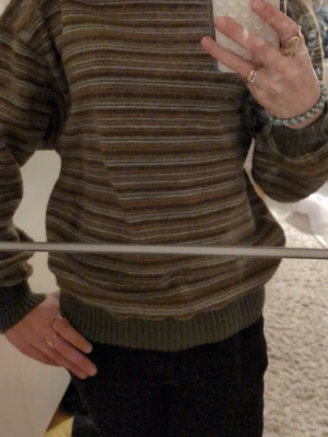 Randig stickad tröja i jordnära toner - Säljer en stickad tröja med horisontella ränder i grönt, brunt och beige. Tröjan har avslappnad passform, ribbade muddar och rund hals. Perfekt för dig som mysiga plagg