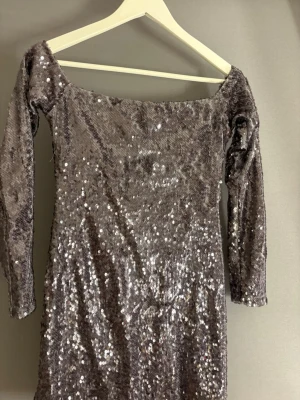 Glittrig långärmad klänning Design by Si - Säljer en glittrig långärmad klänning från Design by Si. Klänningen har en rak, figurnära passform och är täckt av silverfärgade paljetter som ger ett riktigt partyvibe. Offshoulder-modell och kort längd, perfekt för dig som vill sticka ut.