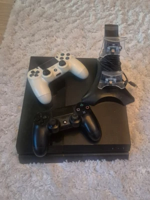 Playstation 4 med två kontroller - Säljer en svart Playstation 4 med en svart och en vit DualShock 4-kontroll samt en laddstation för kontrollerna. Konsolen har en stilren design i plast och passar perfekt för gamingkvällar med vänner.