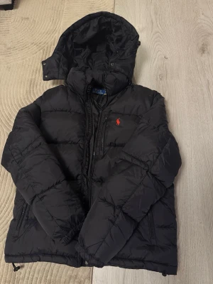 Svart dunjacka från Polo Ralph Lauren - Snygg svart dunjacka från Polo Ralph Lauren med huva och dragkedja. Jackan har det klassiska röda Polo-logot på bröstet och två sidofickor med dragkedja. Perfekt för kalla dagar och riktigt skön att bära.