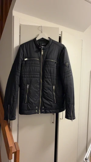Svart vindjacka från Diesel - Snygg svart vindjacka från Diesel med quiltade detaljer och flera dragkedjor framtill. Jackan har hög krage, lång ärm och coola metalldetaljer vid fickor och ärmslut. Perfekt för dig som vill ha en stilren och modern look.