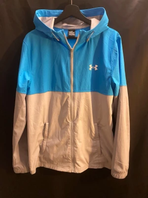 Windbreaker Under Armour - Sällsyn Under Armour vindjacka/windbreaker i blått och grått – en lätt, bekväm och funktionell jacka som skyddar bra mot vind och funkar perfekt både till träning och vardag. Utrustad med huva, hel dragkedja och två fickor för extra komfort och praktisk användning.  Snygg passform, lätt att matcha och i fint skick. Ett riktigt bra kap – passa på innan den försvinner. Kontakta mig vid intresse 👍