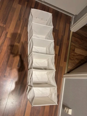 Vit hängande förvaringshylla tyg - Praktisk vit hängande förvaringshylla i tyg med fem fack. Perfekt för garderoben eller hallen för att organisera kläder, skor eller accessoarer. Hyllan har en ögla upptill för enkel upphängning på stång eller krok. Enkel och stilren design.