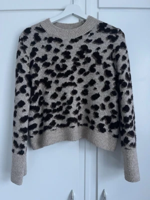 Beige leopardmönstrad stickad tröja - Säljer en beige stickad tröja med leopardmönster i svart och mörkbrunt. Tröjan har rund halsringning, långa ärmar och ribbade muddar vid ärmslut och nederkant. Perfekt för dig som gillar djurmönstrat och vill ha en trendig look.