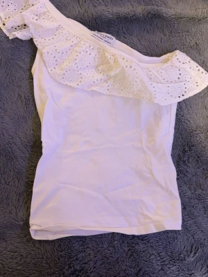 Vit offshoulder topp Holly & White - Söt vit offshoulder topp från Holly & White by Lindex. Toppen har en bred volang med broderade hål och är axelbandslös. Perfekt för sommarens alla tillfällen och ger en romantisk vibe till din outfit.