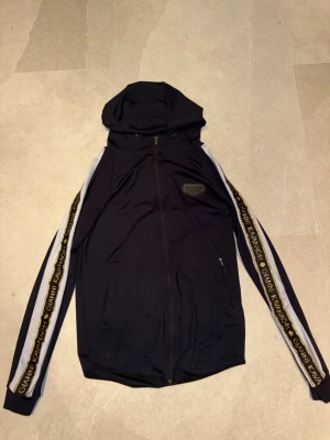 Svart vindjacka Gianni Kavanagh - Svart vindjacka från Gianni Kavanagh med huva och hel dragkedja. Jackan har svarta och vita ärmar med logotypband längs sidorna där det står 'Gianni Kavanagh' i guld. Två fickor framtill och sportig look.