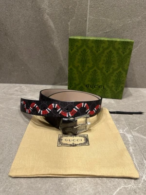 Svart Gucci bälte med ormtryck - Snyggt svart bälte från Gucci med det klassiska GG-mönstret och en rödvit orm som slingrar sig längs bandet. Bältet har en silverfärgad metallspänne och insidan är ljusbeige. Perfekt för att lyfta vilken outfit som helst.