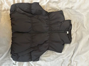 Svart puffig dunväst med hög krage - Säljer en svart dunväst med puffig design och hög krage. Västen har dragkedja framtill och markerade sömmar som ger en cool, oversized look. Perfekt för lager på lager under kyliga dagar. Enkel och stilren utan synligt märke.