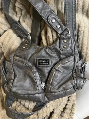 Grå handväska från OUYALU - Cool grå handväska från OUYALU med bred axelrem och flera dragkedjefickor. Väskan har en unik form och detaljer i silverfärgade nitar. Tillverkad i syntetmaterial som ger en skinnliknande look. Perfekt för dig som vill ha en praktisk och snygg accessoar.