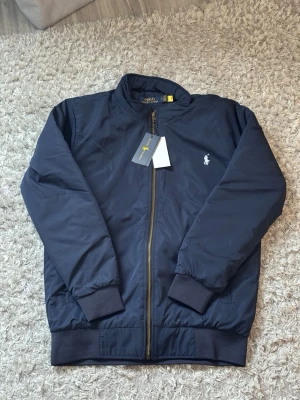 Marinblå bomberjacka från Polo Ralph Lauren - Snygg marinblå bomberjacka från Polo Ralph Lauren med klassisk passform. Jackan har dragkedja framtill, ribbade muddar och den ikoniska vita logon broderad på bröstet. Perfekt för dig som vill ha en stilren och tidlös look.