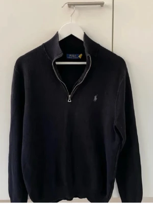 Svart halvzip från Polo Ralph Lauren - Klassisk svart halvzip-tröja från Polo Ralph Lauren med broderad logga på bröstet. Tröjan har hög krage, dragkedja och ribbade muddar. Perfekt för en stilren och avslappnad look.