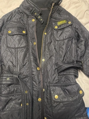 Svart quiltad vindjacka Barbour - Säljer en svart quiltad vindjacka från Barbour International med guldiga knappar, bälte i midjan och flera fickor. Jackan har hög krage, dragkedja och tryckknappar samt ett snyggt Barbour-märke på bröstet. Perfekt för dig som vill ha en klassisk och stilren look. 