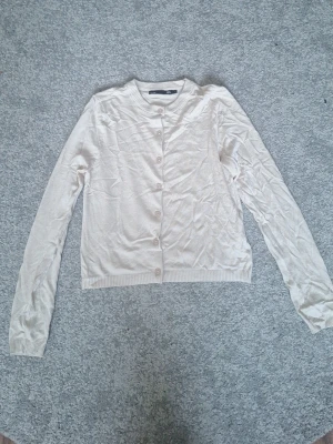 Beige kofta från 157 med knappar - Ljusbeige kofta från 157 med rund halsringning och knäppning framtill. Tillverkad i mjukt, tunt stickat material med ribbade muddar vid ärmslut och nederkant. Perfekt att slänga över en topp för en enkel och clean look. Helt oanvänd och knappar följer med!