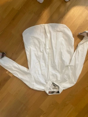 Vit skjorta från Burberry Brit - Klassisk vit skjorta från Burberry Brit i bomull. Skjortan har långa ärmar, knappar framtill och detaljer med Burberrys ikoniska rutmönster på insidan av kragen och manschetterna. Perfekt för en clean och stilren look. Passar XS bäst, men står M på tagen av någon anledning 