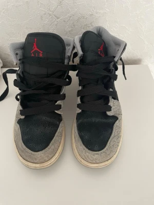 Nike Air Jordan 1 Mid svart/grå - Säljer ett par Nike Air Jordan 1 Mid sneakers i svart och grått med vita detaljer. Skorna har klassisk snörning, svart swoosh och ikoniska Air Jordan-loggan på sidan. Ovandelen är i mocka och skinn med mönstrade grå partier och svart tå.