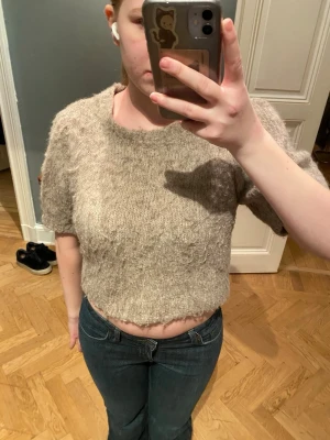 Beige stickad cropped tröja - Mysig beige stickad tröja med kort ärm och croppad passform. Tröjan har en rund halsringning och är stickad i ett luddigt garn som ger en avslappnad look. Perfekt att styla med jeans eller kjol.
