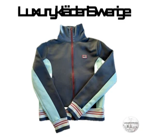 Hysteric Glamour zip - Snygg ziptröja från Hysteric Glamour i mörkblå och ljusblå färg med röda detaljer. Tröjan har lång ärm, högt krage och HG-logga på bröstet. Ränder i rött och vitt på muddar och krage, samt hel dragkedja framtill. Perfekt för dig som gillar retrostil. Strlk S/XS