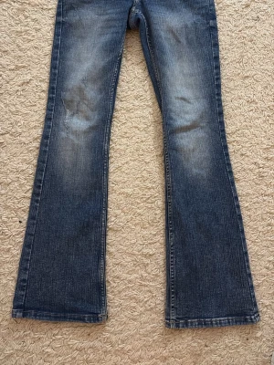 Blå bootcut jeans - Säljer ett par blå bootcut jeans med snygga slitningar på framsidan och klassiska bakfickor med vita sömmar. Jeansen har en lätt utsvängd passform nedtill och är perfekta för en avslappnad och trendig look.