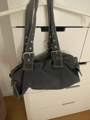 Mörkgrå handväska med spännen - Snygg mörkgrå handväska från clomplement med dubbla axelband och coola metallspännen. Väskan har en mjuk form och är tillverkad i slitstarkt tygmaterial. 