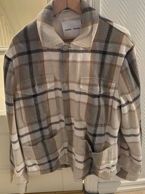 Rutig overshirt från Samsøe Samsøe - Snygg rutig overshirt från Samsøe Samsøe i beige, grå och bruna toner. Overshirten har två bröstfickor, knäppning framtill och en avslappnad krage. Tillverkad i återvunnet material och perfekt för lager på lager-stil.