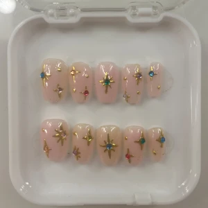 Rosa lösnaglar med stjärndekorationer - Snygga rosa lösnaglar dekorerade med guldstjärnor och färgglada stenar i blått, rött och grönt. Perfekt för en glamorös look.