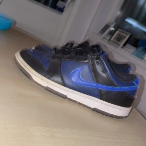 Nike Dunk Low svart/blå sneakers - Nike Dunk Low sneakers i svart och blått med klassisk vit sula. Skorna har svart snörning, rund tå och den ikoniska blå swoosh-loggan på sidan. Ovandelen är i skinn och textil, vilket ger en snygg kontrast mellan färger och material.