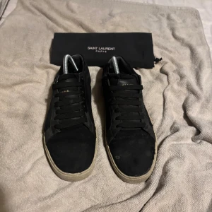 Svarta Saint Laurent sneakers - Svarta sneakers från Saint Laurent med vit sula och svart snörning. Sidan pryds av Saint Laurent-logga i vitt och insidan har guldfärgad text. Ovandelen är i mocka och skinn, med klassisk låg profil och rund tå. Skorna levereras med originalpåse. OBS! Litet hål bak på skon. Varan tvättas självklart innan den säljs. Skriv för fler funderingar. Dustbag kommer med