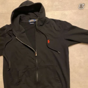Svart hoodie från Polo Ralph Lauren - Svart hoodie från Polo Ralph Lauren med röd broderad logga på bröstet. Tröjan har dragkedja, huva med snörning och två stora fickor framtill. Tillverkad i mjuk bomull och har lång ärm. Perfekt för en chill och stilren look.