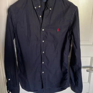 Mörkblå skjorta Polo Ralph Lauren M - Snygg mörkblå skjorta från Polo Ralph Lauren i slim fit-modell. Skjortan har långa ärmar, klassisk krage med knappar och röd broderad logga på bröstet. Tillverkad i bomull och passar perfekt till en stilren look.
