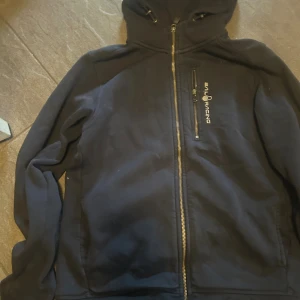 Svart hoodie från Sail Racing med fleece - Svart hoodie från Sail Racing med dragkedja hela vägen, broderad logga på bröstet och praktisk bröstficka med dragkedja. Insidan är fodrad med mjuk fleece och huvan har snörning. Perfekt för kyliga dagar och snygg till jeans.