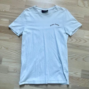 Vit t-shirt Closure London XS - Vit t-shirt från Closure London i storlek XS. Framsidan har en liten svart logga och baksidan har ett stort tryck med Palm Springs-motiv i lila och blå toner. Klassisk rund hals och korta ärmar. Mjuk bomullskvalitet, perfekt för chill dagar.